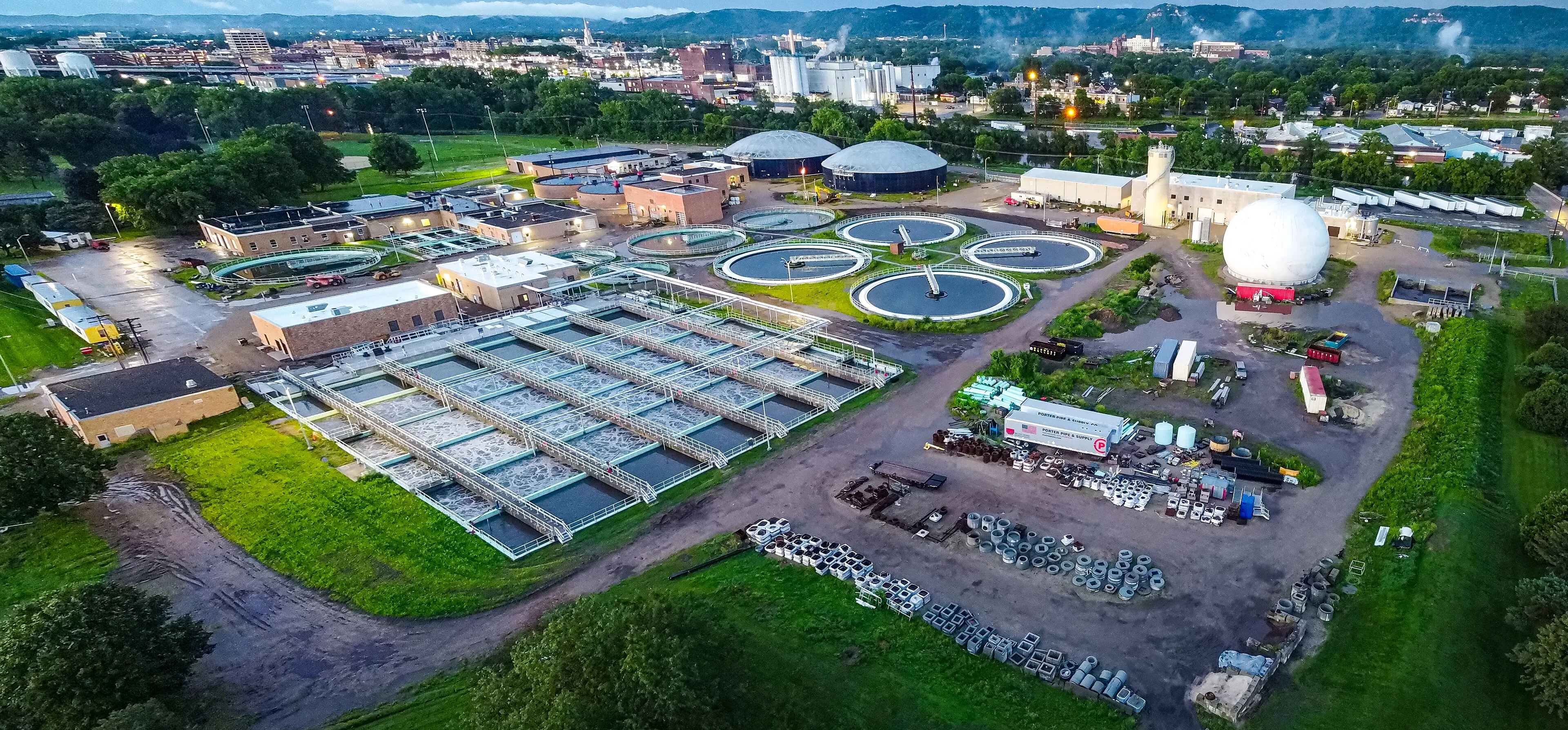 Aerial view of a wastewater treatment facility at dusk, with circular clarifiers and storage domes set against a city skyline and distant hills.